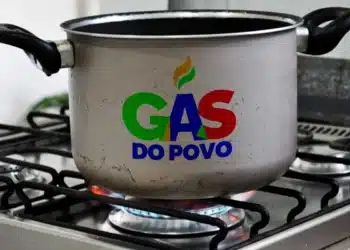 Panela de alumínio sobre fogão a gás com chama azul e logo colorido do programa Gás do Povo.