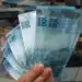 Mão segurando leque de notas de R$ 100 com maços de dinheiro ao fundo