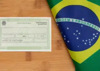 Título de eleitor brasileiro com identificação biométrica ao lado da bandeira do Brasil sobre superfície de madeira.