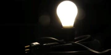Lâmpada incandescente acesa em destaque sobre um fundo escuro, com um cabo de energia e plugue preto em primeiro plano.