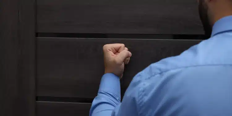 Homem vestindo camisa social azul bate com a mão fechada em uma porta de madeira escura, visto de costas.