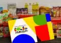 Cartão colorido do Programa Bolsa Família sobre superfície escura, com garrafas de óleo, pacotes e produtos ao fundo