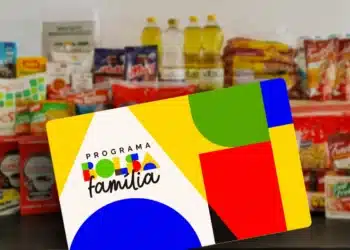Cartão colorido do Programa Bolsa Família sobre superfície escura, com garrafas de óleo, pacotes e produtos ao fundo