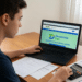 Jovem de camiseta azul usa notebook para acessar o site Desenrola Fies, com calculadora e documentos sobre mesa de madeira
