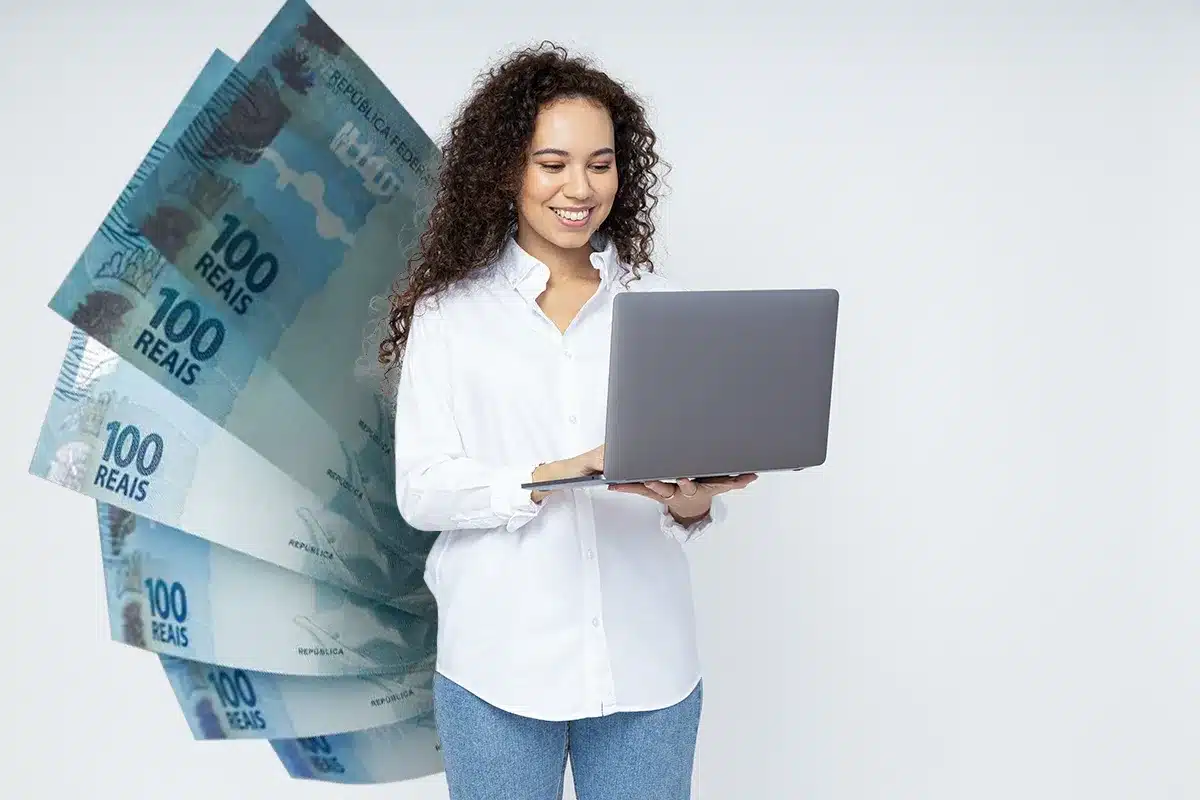 Mulher de cabelos cacheados usando camisa branca segura laptop cinza com notas de 100 reais em leque ao lado