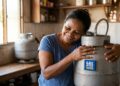 Mulher sorridente de camiseta azul abraça botijão de gás Ultragaz com adesivo Gás do Povo 2026 em cozinha humilde