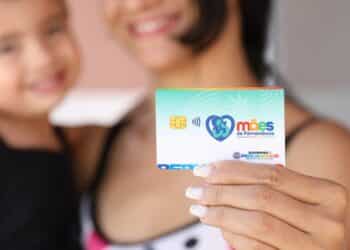 Mãe segura cartão Mães de Pernambuco com chip e logo do governo estadual, criança ao fundo desfocada