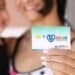 Mãe segura cartão Mães de Pernambuco com chip e logo do governo estadual, criança ao fundo desfocada