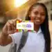 Jovem estudante negra com uniforme branco e mochila mostrando cartão do Pé-de-Meia