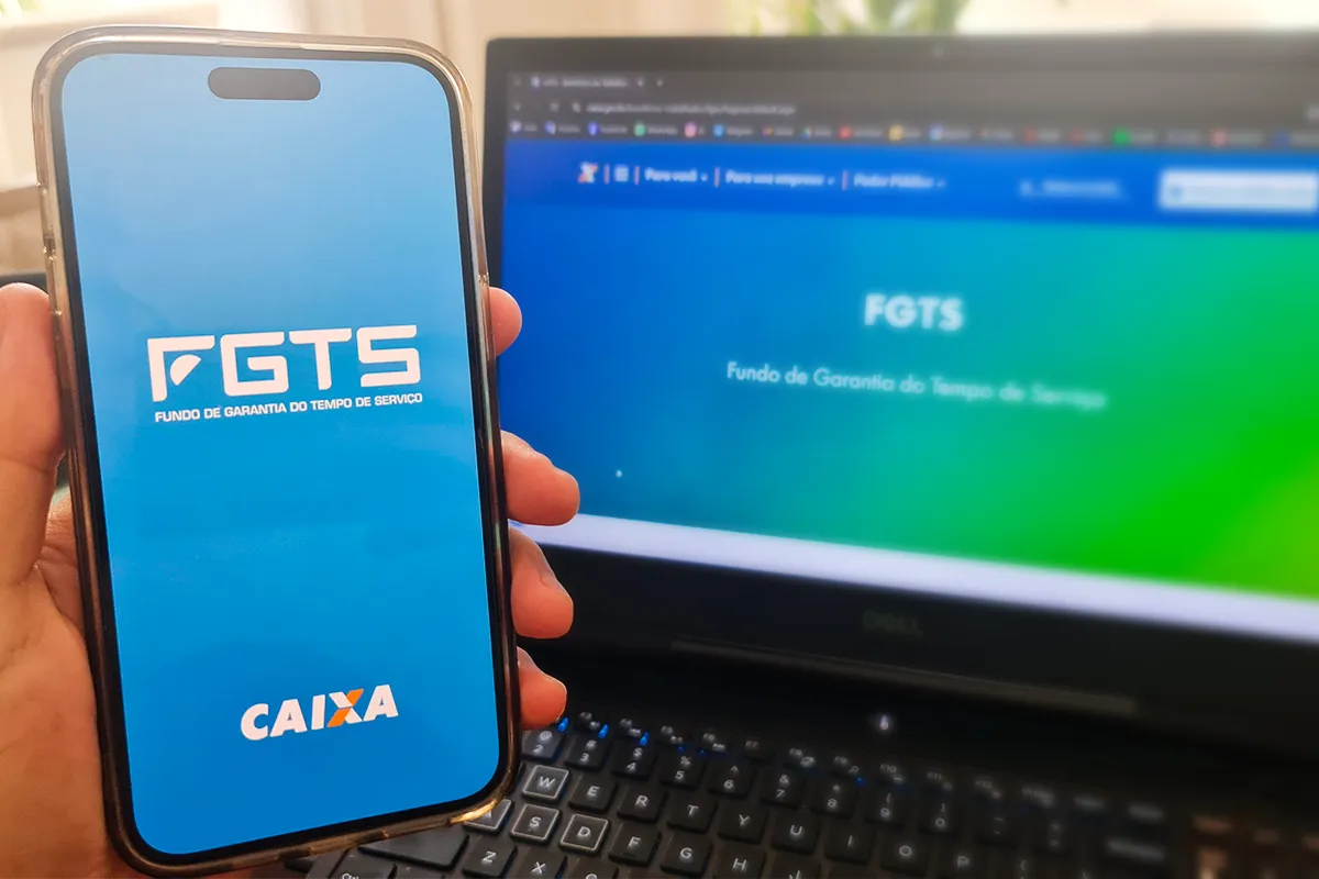 Mão segura smartphone exibindo tela azul do aplicativo FGTS com logo da Caixa; notebook ao fundo mostra portal FGTS
