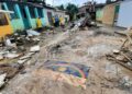 Rua destruída com escombros, lama e bandeira de Pernambuco no chão; equipes de resgate ao fundo