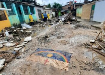 Rua destruída com escombros, lama e bandeira de Pernambuco no chão; equipes de resgate ao fundo
