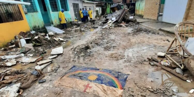 Rua destruída com escombros, lama e bandeira de Pernambuco no chão; equipes de resgate ao fundo