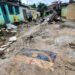 Rua destruída com escombros, lama e bandeira de Pernambuco no chão; equipes de resgate ao fundo