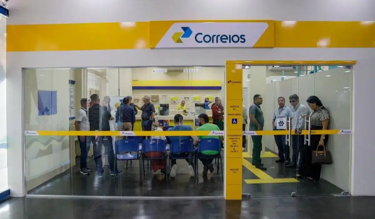 Fachada de agência dos Correios com clientes aguardando atendimento presencial em guichês com cadeiras azuis