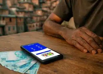Mãos apoiadas em mesa de madeira com celular exibindo aplicativo Bolsa Família e cédulas de R$ 100