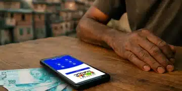 Mãos apoiadas em mesa de madeira com celular exibindo aplicativo Bolsa Família e cédulas de R$ 100