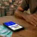 Mãos apoiadas em mesa de madeira com celular exibindo aplicativo Bolsa Família e cédulas de R$ 100