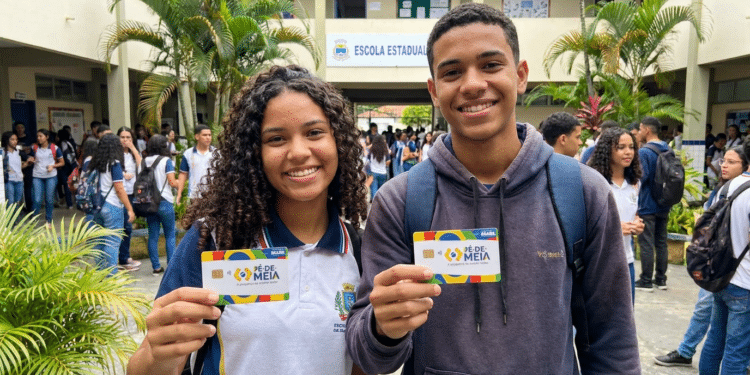 Dois estudantes sorrindo mostram cartões do Pé-de-Meia em frente a escola estadual, com outros alunos ao fundo