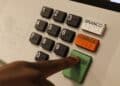 Mão pressionando tecla verde Confirma em urna eletrônica com teclado numérico, botões Branco e Corrige