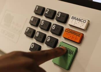 Mão pressionando tecla verde Confirma em urna eletrônica com teclado numérico, botões Branco e Corrige