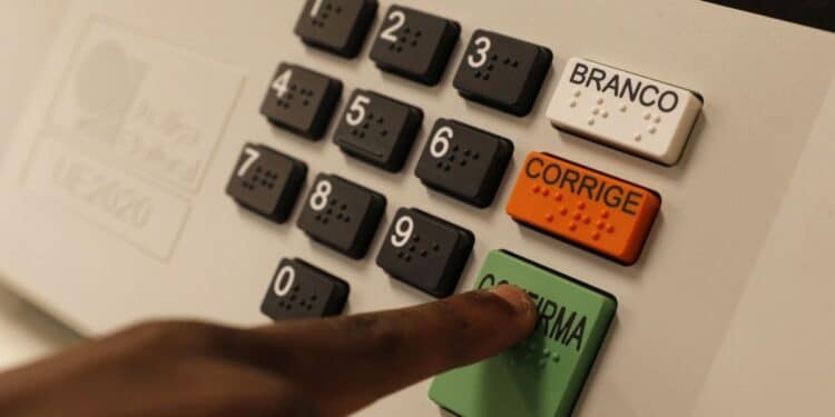 Mão pressionando tecla verde Confirma em urna eletrônica com teclado numérico, botões Branco e Corrige