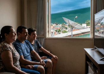 Família assiste à TV em casa enquanto antena parabólica aparece pela janela