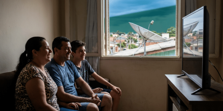 Família assiste à TV em casa enquanto antena parabólica aparece pela janela