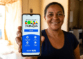 Mulher sorridente mostra celular com tela do app Bolsa Família exibindo menu de acesso e logos da CAIXA e Brasil
