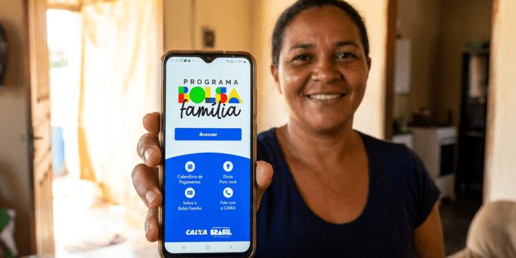 Mulher sorridente mostra celular com tela do app Bolsa Família exibindo menu de acesso e logos da CAIXA e Brasil
