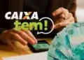 Mãos manuseiam celular com aplicativo Caixa Tem ao lado de notas de R$ 100