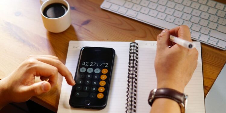 Pessoa usando calculadora no celular enquanto anota valores em um caderno