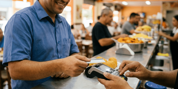 Homem sorridente de camisa azul aproxima cartão de maquininha em restaurante self-service com pratos de comida ao fundo