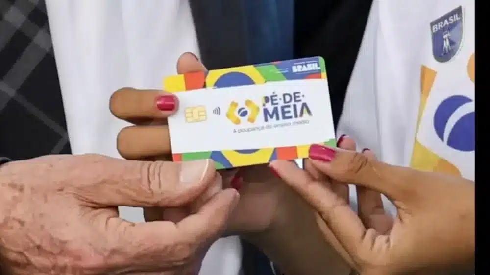 Mãos segurando cartão Pé-de-Meia com chip dourado, logotipo colorido e emblema do MEC ao fundo