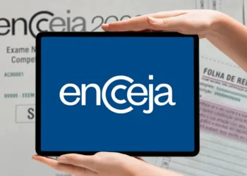 Logo do Encceja exibido em um tablet segurado por duas mãos, com documentos de prova e folha de redação ao fundo.