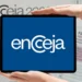 Logo do Encceja exibido em um tablet segurado por duas mãos, com documentos de prova e folha de redação ao fundo.