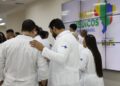 Profissionais de jaleco branco com logo SUS reunidos em evento com painel Mais Médicos para o Brasil ao fundo