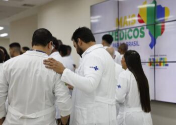 Profissionais de jaleco branco com logo SUS reunidos em evento com painel Mais Médicos para o Brasil ao fundo