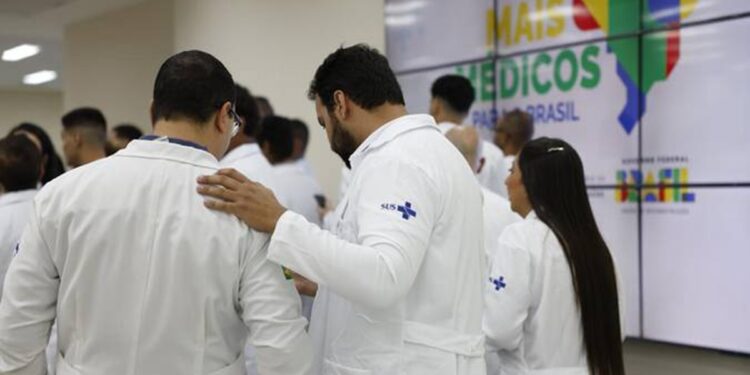 Profissionais de jaleco branco com logo SUS reunidos em evento com painel Mais Médicos para o Brasil ao fundo