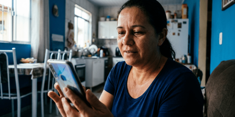 Mulher de cabelos escuros presos, camiseta azul, sentada em sofá consultando celular em ambiente doméstico