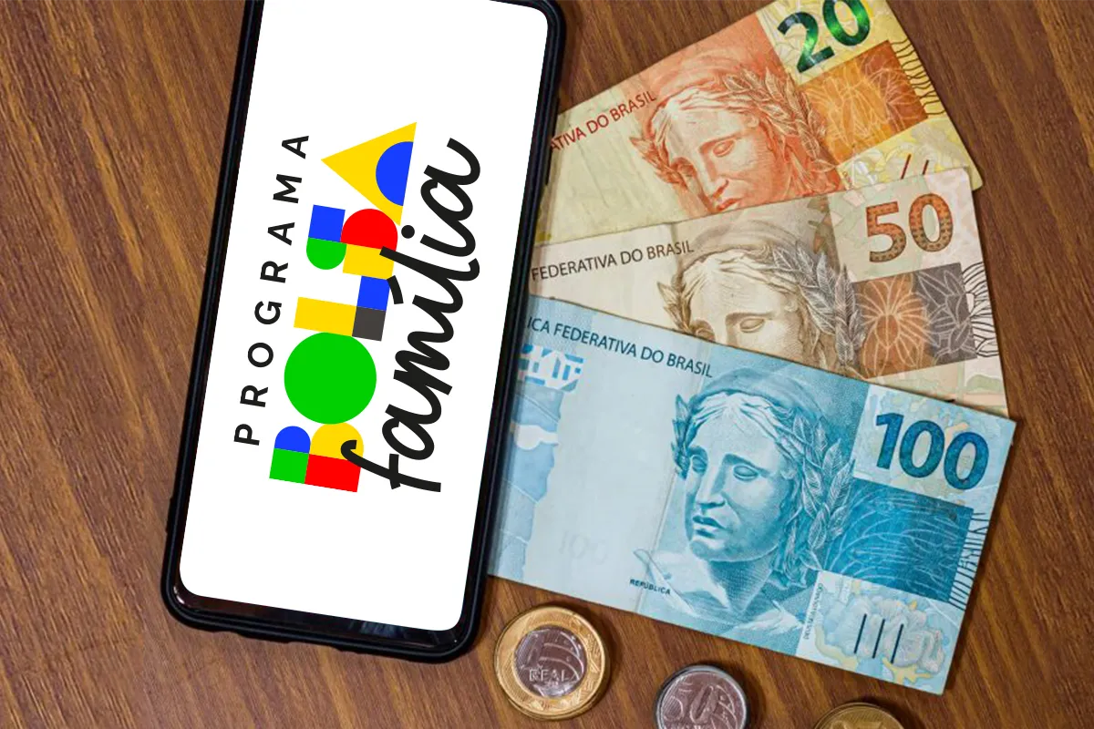 Smartphone sobre mesa de madeira exibindo o logotipo do Programa Bolsa Família, acompanhado de cédulas de 20, 50 e 100 reais e moedas de real.
