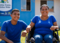 Mulher em cadeira de rodas e jovem sorrindo em frente a casa azul do programa Minha Casa, Minha Vida