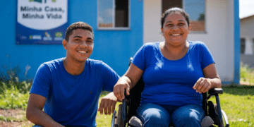 Mulher em cadeira de rodas e jovem sorrindo em frente a casa azul do programa Minha Casa, Minha Vida