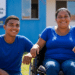 Mulher em cadeira de rodas e jovem sorrindo em frente a casa azul do programa Minha Casa, Minha Vida