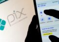 Pessoa utilizando aplicativo bancário no celular para realizar um PIX, com o logotipo do PIX em destaque ao fundo em uma tela de computador.