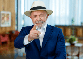 Presidente Lula vestindo paletó azul e chapéu panamá claro, apontando o dedo indicador para a frente em um gesto de ênfase, em um ambiente interno iluminado.