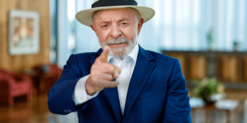 Presidente Lula vestindo paletó azul e chapéu panamá claro, apontando o dedo indicador para a frente em um gesto de ênfase, em um ambiente interno iluminado.