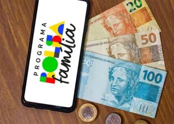 Um smartphone sobre uma mesa de madeira exibe o logotipo do Programa Bolsa Família. Ao lado do aparelho, há cédulas de 20, 50 e 100 reais, além de moedas de um real e 50 centavos.