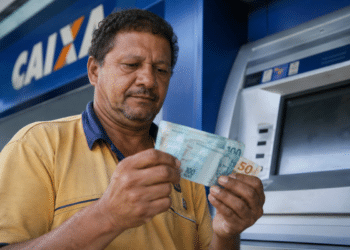 Homem de camisa amarela confere notas de R$ 100 e R$ 50 em frente a um caixa eletrônico da Caixa