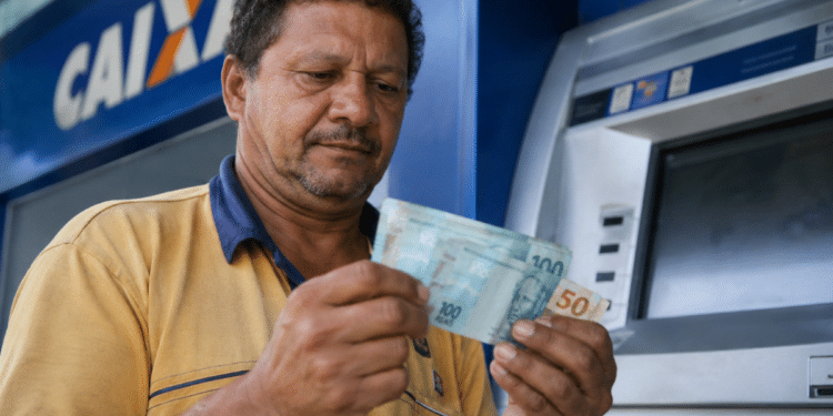 Homem de camisa amarela confere notas de R$ 100 e R$ 50 em frente a um caixa eletrônico da Caixa